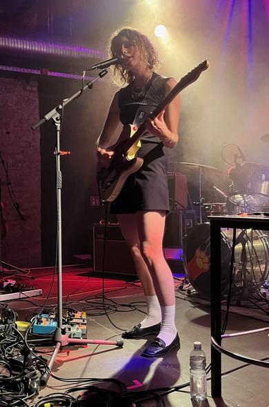 lead singer alone on stage playing guitar and singing;spot lights surrounding her are red and purple, on the floor a shadow is visible