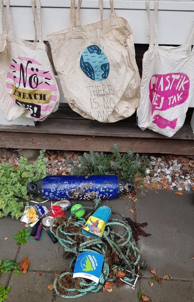 Gesammelter Strandmüll.
Im Hintergrund 3 waschbare Stoffbeutel, in denen ich den Müll sammle, den ich tragen kann.
 Im Vordergrund ein PlastikfreeHindsholm-Aufkleber.

Auf dem ersten Stoffbeutel der Aufdruck: Nö Einfach Nö!
2ter Aufdruck: There is no planet B
3ter Aufdruck: Plastik ne tak