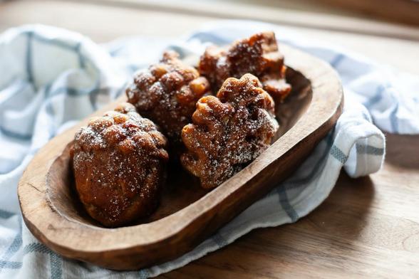 A tightly cropped horizontal image of a collection of four powder sugar dusted caramelly brown baked goods in the shapes of acorns and leaves. They're arranged in a narrow rough-carved wooden bowl at a diagonal angle to the camera. The bowl is sitting on a rumpled tea towel of pale blue stripes, on a wooden cutting board.