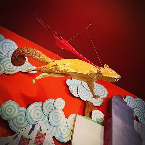 Looking up at a detailed squirrel fashioned of cardboard. It’s paws and tail are outstretched, flying, and a red cape around its neck flows backward. Cardboard clouds and cityscape behind it.