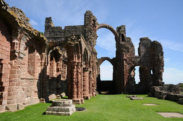 Lindisfarne Priory. The image shows a view along the ruined priory church with a ruined red stone wall on the left and remains of taller structures and a high arch slightly further into the frame. The ground is covered with grass and there are traces of columns in the foreground and on the right, with more complete ones further away. The scene is in sunlight.