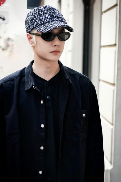 Wang Yibo at Chanel flagship store in Paris. he has platinum blond hair under a tweedy baseball cap. he's wearing black sunglasses and a rumpled, oversized black shirt.