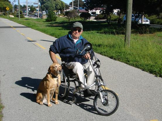 A man is riding a hand cycle on a paved, car-free multiuse trail in Monterey. He is accompanied by a leashed dog, his golden companion! The man is wearing sunglasses, a dark grey baseball cap, light grey pants, white shirt, and dark blue jacket with turquoise lining. No one else is visible on the trail, and to the right, separated by a green belt,  there are motor vehicles on the street, Del Monte Avenue.