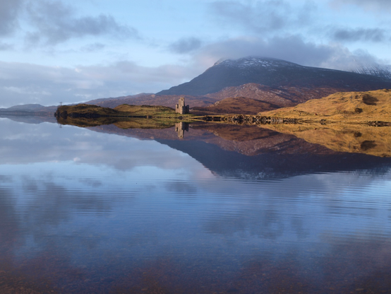 A photo of a loch with ruined castle.  Blue sky with great white clouds, a mountain centre right with a hint of snow on the peak, inform a ruined castle on a promontory into a calm Loch which reflects the sky, the hill, the castle, A raised land humo to the far right green golden in the sun.