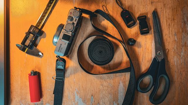 A wooden table. On it a collection of objects.
Lighter, digital caliper, Fuji X70, quick release, buckle, a 1/4" eyelet, scissors, nylon belt.
The X70 is already attached to a nylon strap with quick-release buckles at the lugs. Some parts of the nylon strap are fixed with paper clips.