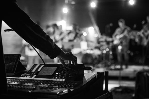 A black and white photo of a Yamaha LS9 digital mixer with a band playing in the background.
