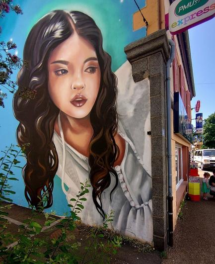 Streetartwall. On an exterior wall on a house wall on a street corner was sprayed / painted beautiful mural of a young woman. The background is designed as light blue color arcs with small bright stars. The girl has long brown and wavy hair. Her face is bright and finely drawn. She wears a white dress, maybe a nightgown, with small buttons. She is looking curiously toward the street. (The photo shows a commercial street with a tobacco/lottery store, in front of which a child with mother is standing).