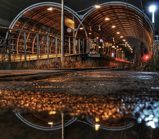 Eine Haltestelle der Stadtbahn Bonn bei Nacht spiegelt sich zun Teil in einer Pfütze. Die Haltestelle ist von zwei halbrunden Stahlkonstruktionen überdacht, die von innen beleuchtet werden.