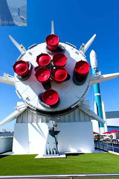An outdoor, daylight photograph of a white, circle with four red cones on the outside, and four in the center and with six small fins on the circumference. It is held up by a metal brace. Behind it is a blue and white rocket and the lightly cloudy blue sky.