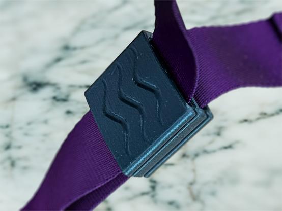 A black 3d printed webbing buckle, mounted on violet webbing in front of a marble surface.