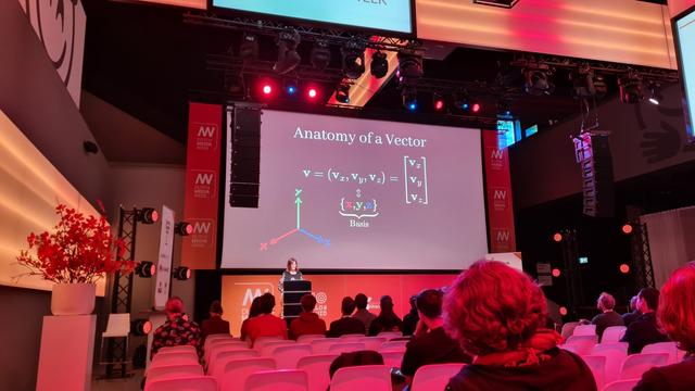 Freya standing behind a lectern on a stage, with an audience. The slides projected on screen read "anatomy of a vector".