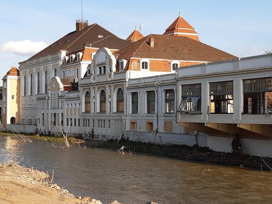 Dasselbe Gebäude nach der Flut. Man erkennt, dass die Fenster zerstört sind, auch der Uferbereich weist deutliche Schäden auf.