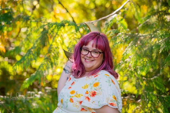 A pink-haired, fat white woman in a summer dress smiles in a sunny forest, holding a brass moon behind her head.