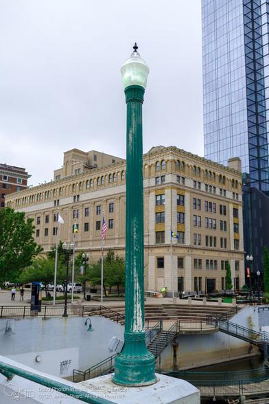 An antique-style green lightpole with a fluted column shape emerges from a concrete bridge pier. Behind it a circular set of cement seats descends to stairs and finally to a riverside dock and boardwalk. An 8-story yellow brick and terra-cotta building sits beyond with arched windows and decorative details on the top floor. The glass wall of modern skyscraper ascends out of the top right edge of the frame.