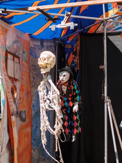A color photograph of two puppets - marionettes -hanging from a stand backstage between shows. The leftmost puppet is a skeleton with bloodshot eyes in a skull, the rest of the “body” made from a white wooden clothes hanger with loops of white fabric strung below, the rightmost puppet is a somewhat sinister looking clown with a pointed face, wearing dark clothing with a harlequin pattern. The brightly colored tent fabric around them is lit by sunlight.