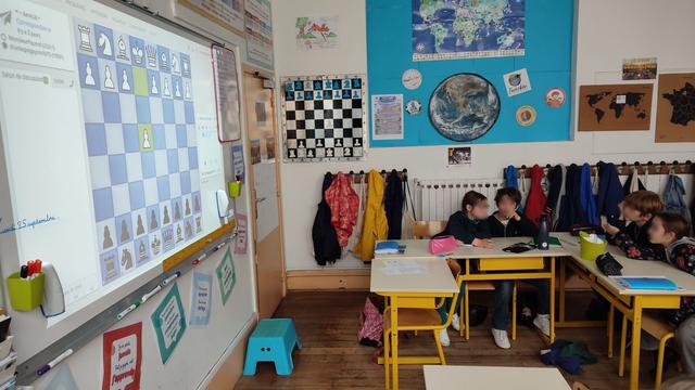 Une salle de classe où l'on voit des élèves et un écran numérique avec la partie d'échecs en cours.