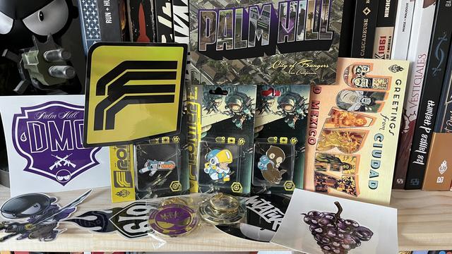 Ensemble de produits de la boutique du label 619 : trois pins issus de la BD Frontier de Singelin, et deux portes clefs jeton de casino en métal issu de Mutafukaz 2 (tome 2). Autour, plusieurs stickers : le logo de Frontier, la tête de Lino (Mutafukaz), Lino avec un katana, une grappe de raisin ou la tête de Lino remplace les raisins et le logo 619. Deux cartes postales sont également visibles, de Palm Hill et Juaréz.