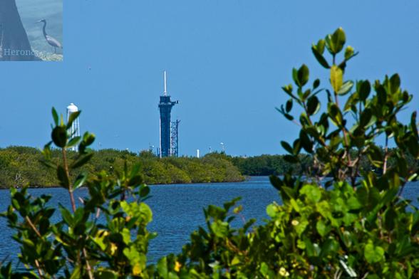 An outdoor photograph of Launch Complex 39A with the Banana River in the foreground, as well as some scrub bushes. On the other shore is more green scrub land.  Further in the distance a black tower rises with a tall, white cylinder on its top, which provides access for the crew on the Crew Dragon spacecraft.  There is a large water tower on the left side of the image.