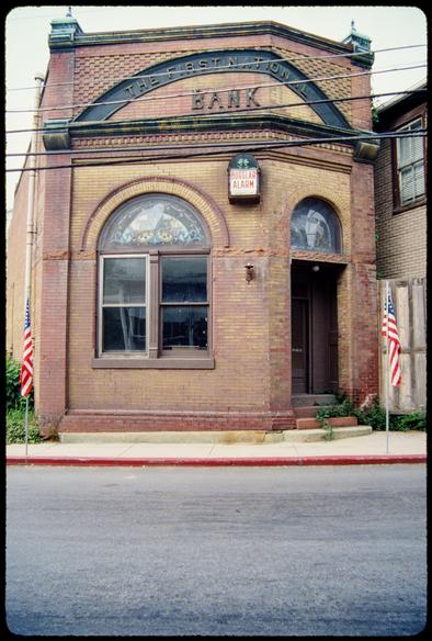 A small one-story bank building, brick with a few stone details, stands along a sidewalk on an empty street. The building is just wide enough for an entry door and two side-by-side windows topped by an arched stained glass window. At the top, a sign reads “The First National” in an arch over the larger word “Bank.” A somewhat large and out-of-scale Burglar Alarm box (so labelled) is attached to the front of the building. A pair of U.S. flags stand on short pole along the sidewalk.