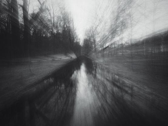 Black and white photo  of a stream of water, river like, in the centre of the photo and trees on the sides but the zoom lens was zoomed out giving a warp effect to the photo.
This is somewhere in Dresden, Germany.