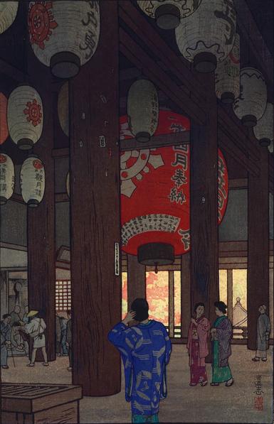 A view of visitors in the interior of Ishiyama-dera on an autumn day. In the foreground, a female figure in a blue-patterned kimono faces away from the viewer, possibly looking at the enormous red lantern suspended from the temple ceiling. Other visitors to the temple go about their business: a pair of female figures in kimono, a male figure in a small hat looking up at the red lantern, a male figure leaning on a cane, a figure wearing the traditional white robes and wide straw hat of a pilgrim, and several more. The many lanterns suspended from the temple ceiling are unlit, and the atmosphere is shaded and dim. Beyond the temple, bright sunshine makes the autumn colours marking the trees seem more intense.
