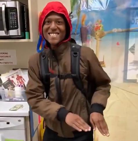 Photograph of Elijah McClain wearing a brown jacket, red hood and a black knapsack in the break room at his place of employment. He's depicted smiling very pleasantly as he seems to be demonstrating dance moves.