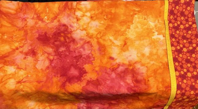 A handmade pillowcase is draped over an ironing board. The main body of the pillow is red and orange ice-dyed, the cuff is a red, orange and yellow pattern of circles, and the trim is a fun yellow with some texture.