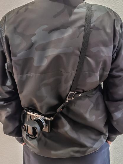 Person wearing a three point camera strap over a camouflage jacket. Seen from the back.