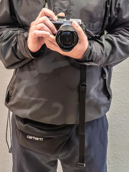 Person wearing a three point camera strap over a camouflage jacket. Seen from the front. The strap cross strap is now disengaged and person is holding the camera to shoot.