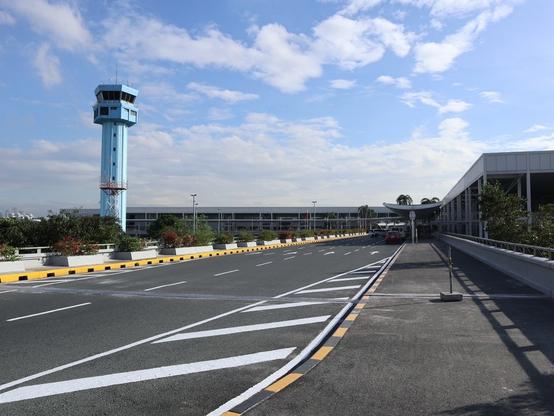 The image features an airport scene with a blue control tower visible in the background. A few cars are parked closer to the control tower, while others are scattered around the area. 

NAIA 2. The NAIA 2 Terminal