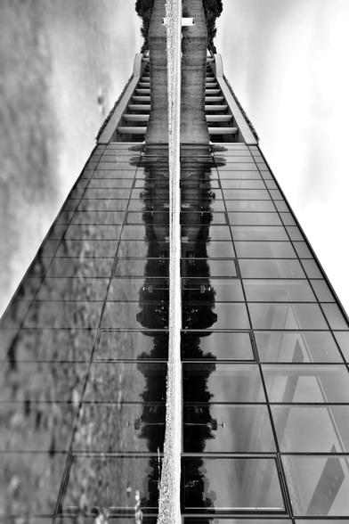 Um 90 Grad gedrehtes SW-Foto mit der Pfützenreflexion eines flachen Gebäudes aus Glas, das von der Seite aus fotografiert wurde, sodass es so aussieht, als ob sich das Gebäude samt Spiegelung nach oben verjüngt. In der Glasfassade spiegeln sich Bäume und der bewölkte Himmel - was sich wiederum in der Pfütze spiegelt.