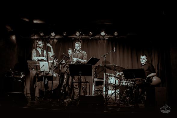 Schwarz-weiß Bild der Band Bass 'n Two auf der Bühne. Links ein Kontrabass Spieler, dahinter der Gitarrist, im Vordergrund hinter Notenständern der Saxophonist; weiter rechts der Schlagzeuger.