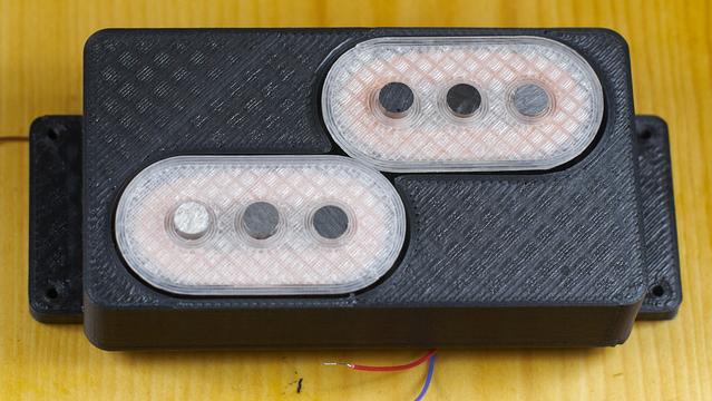 A guitar pickup in the large fender widerange size. There are two bobbins with 3 5mm alnico magnet polepieces each arranged in a staggered arrangement within the frame made to fit the widerange size. The bobbins are 3D printed from natural translucent PLA and the frame or housing is printed from black PLA with carbon fiber. The printed parts show a checked pattern from the infill pattern used. The copper coil windings are dimly visible through the translucent natural PLA.