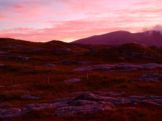 A sunset photo.A salmon pink sky with graduates pink clouds with hills below silhouette pd. A rocky moorland landscape in the foreground wip high is glowing pinky orange with the sun.