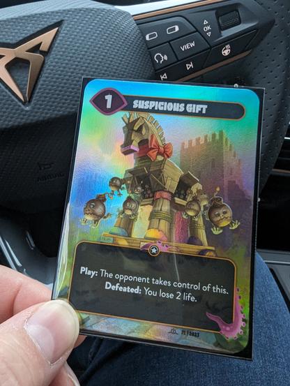 A card from a card game. Upper left shows 1 as its strength. Card name on top: suspicious gift.
In the bottom its effect: 

Play: The opponent takes control of this. Defeated: You lose 2 life.

The artwork shows a big wooden horse, a stone tower in the background. Out of several openings, creatures drop that look like bombs.

The card is a "shiny", with colorful background