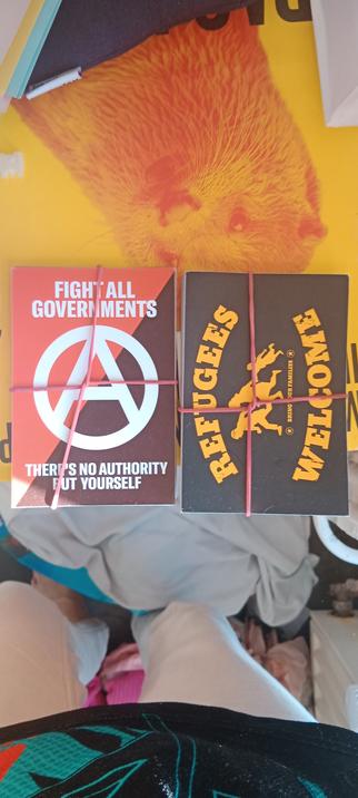 2 heap of stickers. 
The one on the left is a white "A" suround by a circle and with a red and black background like the anacho comunist flag. It's written "Fight all gouvernements. There is no authority but yourself."
The one on the right are black stickers with a yellow family running. It's written in yellow "Refuges welcome" and in a smaller font "And bring your family"