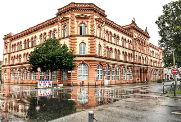 Ein orange-beiges historisches dreistöckiges Gebäude an einer Straßenkreuzung mit vielen gewölbten Fenstern. Die Ecke des Gebäudes sieht wie ein großer Erker oder ein kleiner eckiger Turm aus. Die Kreuzung ist in großen Teilen von Wasser überzogen, sodass sich das Gebäude sowie eine Absperrung und ein Verkehrsschild darin spiegeln. Es ist ein trüber Tag.
