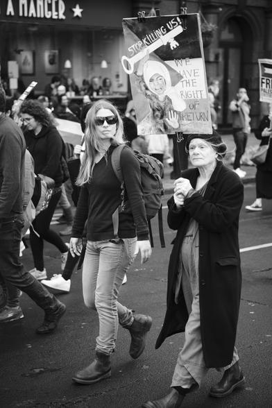 A black and white photograph of one old and one young woman walking in pro-Palestine protest holding a sign reading, "Stop killing us!"