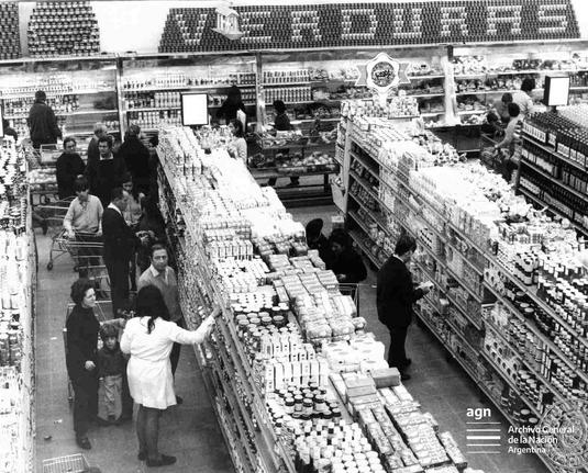 Foto en blanco y negro en la cual se ve, desde arriba, el interior de un supermercado con sus góndolas. 

Gente recorriendo los pasillos con carritos.

Al fondo, en grande, un letrero de “verduras” hecho con cientos de latas de conserva apiladas prolijamente.