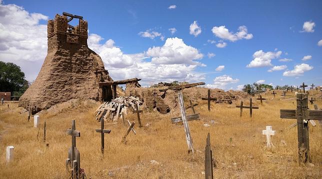 This is a church ruin and cemetery in northern New Mexico.  The remains of the church are made of adobe and graves are marked with simple crosses.  The ground is very tan and the sky is blue.