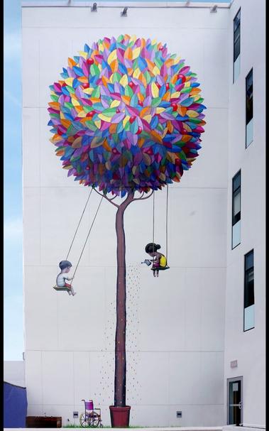 Streetartwall. A mural showing two children swinging on a colorful tree was sprayed/painted on the exterior wall of an approximately four-story modern hospital building. The mural is designed on a white background in the style of naive painting. The tree stands in a clay pot, has a long trunk and its foliage is circular with small leaves in rainbow colors. Below it, a boy is sitting on the left and a girl on the right on small wooden swings. The girl is pouring colorful water on the trunk with a watering can and under the boy is a wheelchair on the ground in the grass.