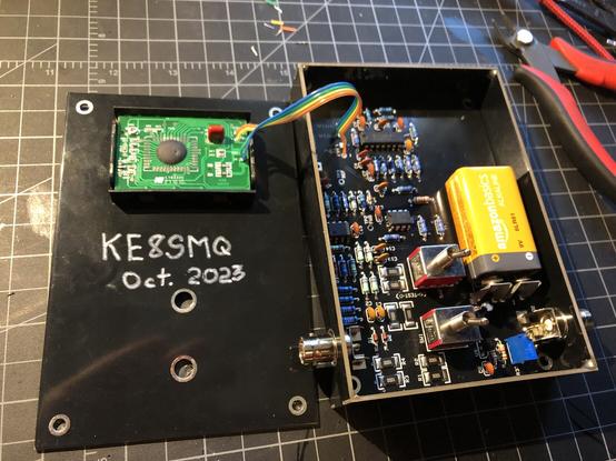A partially assembled power meter kit.

On the left, the back of the top cover, showing the back of the digital display, the callsign "KE8SMQ", and a date of "October, 2023".

On the right, a circuit board with many resistors, capacitors, integrated circuits, two switches, and a battery.