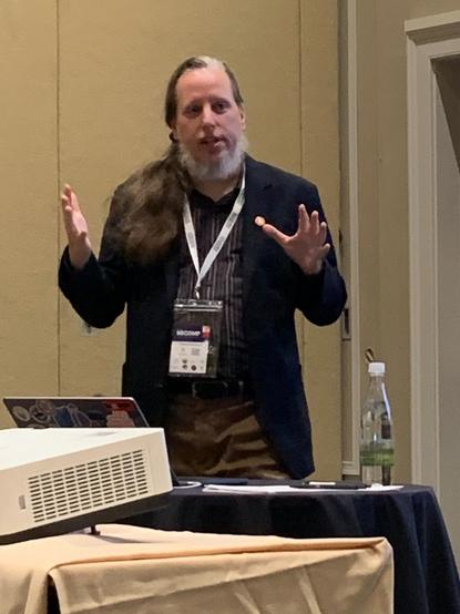 A photo of me wearing a sports coat and a conference pass, doing a presentation in a beige room, holding my arms out like the History-Channel-aliens guy