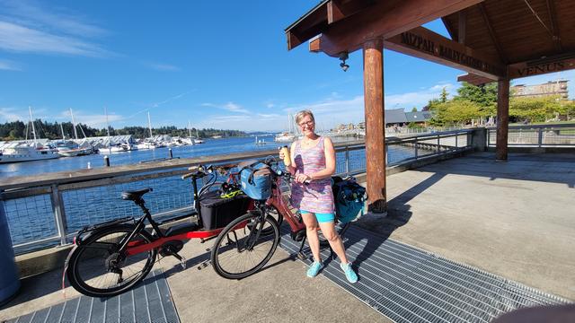 Photo of a white woman with short blonde hair wearing a pastel rainbow tank top dress and turquoise shorts holding a wooden coffee travel cup standing by 3 bicycles parked together on a landing in front of blue water with sailboats in the background
