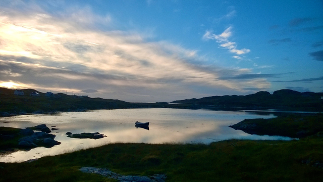 A photo of Loch na Beiste, Harris. Early morning, still not light. Clouds banked to the  left of the shot, a blue sky creeping in on the right. Silhouetted rocky shore line on the horizon giving way to s slither of sea. A calm loch, silver with the cloud reflections, a row boat in silhouette centre shot. Rocks to the left and a grassy bank in the foreground, almost in silhouette but clearly visible.