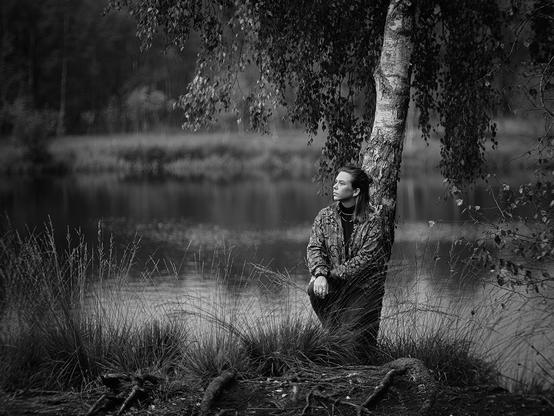 Junge Frau, die an einen Baum gelehnt ist, der direkt an einem See steht.