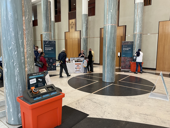 An open foyer with green coloured stone columns and black and white stone flooring. There are some banners and displays with people gathered around.