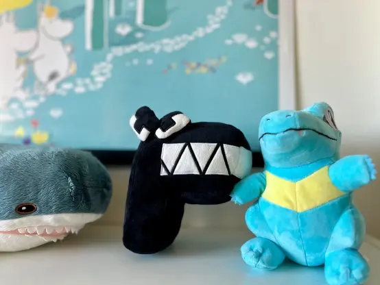 The top of a bookshelf, on top of which lay a number of plushies, the focus of the image is on the one in the center. It's a pitch black character shaped like the capitalized letter "F". This character is from the very popular web series called "Alphabet Lore". The character is designed to look cartoony, and simplistic. The gap between the two "shelves" in the characters' "F" shape is comprised of a sharp set of jagged teeth. He is wearing an angry expression on his face. To his left is a small Blåhaj plushie from IKEA, and to his right, a Totodile plushie from the Pokémon franchise.
The backdrop consists of a white wall, fronted by a summertime themed Moomin poster.