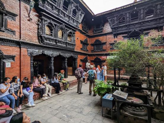 This is a three-story-tall courtyard.  The walls are red stone and the wooden bits around windows and doors are painted black.  There are people sitting on ledges and a small tree in the middle of the courtyard.