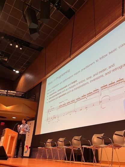 A slide that says "Music == Programming". Devin is at left, speaking at a podium.